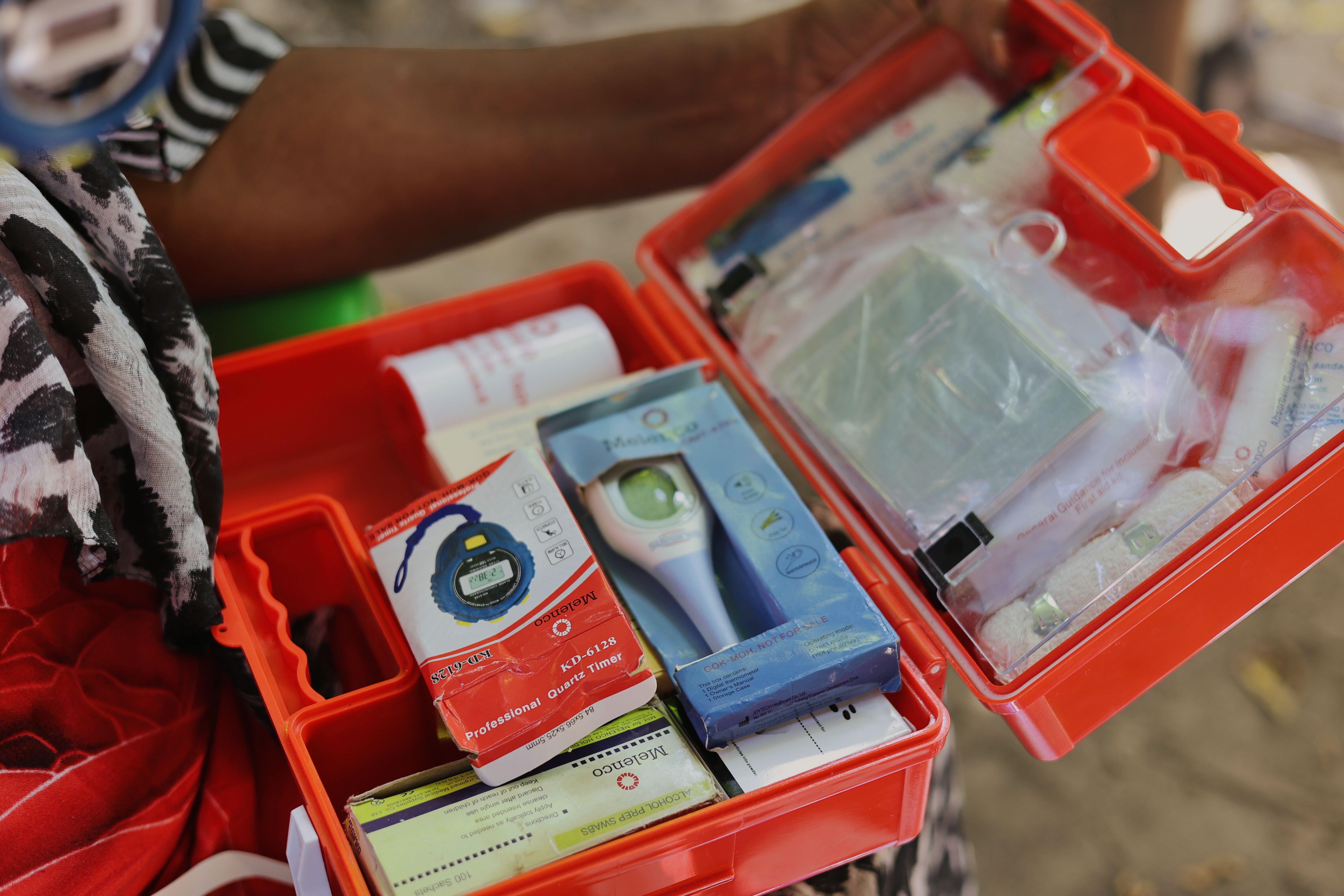Close-up of an open orange first aid kit held by community health worker Mwanamkasi Hassan in Kilifi County, Kenya, containing a boxed digital thermometer, a professional quartz timer, alcohol prep swabs and sterile dressings.